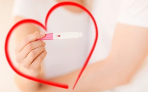 Κοντινή φωτογραφία μιας γυναίκας που κρατά ένα θετικό τεστ εγκυμοσύνης με φόντο μια κόκκινη καρδιά.