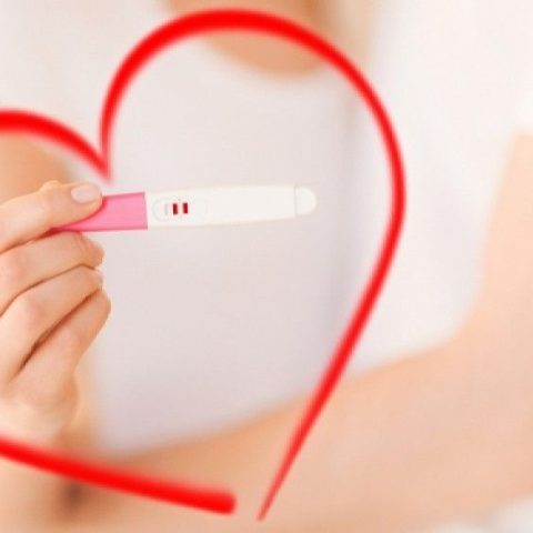Κοντινή φωτογραφία μιας γυναίκας που κρατά ένα θετικό τεστ εγκυμοσύνης με φόντο μια κόκκινη καρδιά.